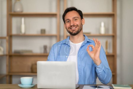 Ein fröhlicher Mann sitzt in einem lässigen Büro, gibt mit der Hand ein positives Okay und blickt den Betrachter direkt an. Er trägt ein blaues Hemd und sitzt vor einem Laptop