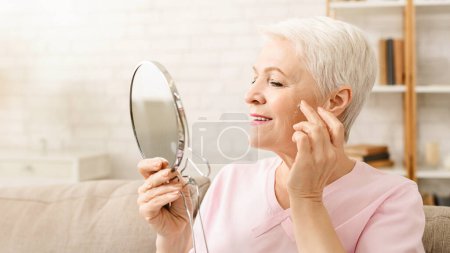 Senior woman holding hand mirror, examining her face closely. She appears to be focused and contemplative as she observes her features intently.
