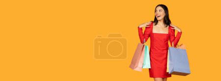 Téléchargez les photos : Une jeune femme avec une expression joyeuse porte plusieurs sacs à provisions alors qu'elle est vêtue d'une robe rouge élégante. Elle semble être à mi-chemin, peut-être à pied, panorama avec espace de copie - en image libre de droit