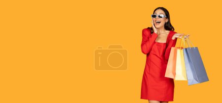 Téléchargez les photos : A woman dressed in a vibrant red dress stands holding multiple shopping bags in her hands, showcasing a successful shopping trip, looking at copy space - en image libre de droit