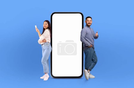 The scene features a young Asian couple, both grinning, positioned next to a life-sized smartphone mockup on a seamless blue background.