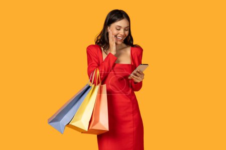 Eine Frau in einem leuchtend roten Kleid beschäftigt sich mit ihrem Smartphone, während sie mehrere Einkaufstüten in den Händen hält. Sie scheint in ihr Gerät vertieft, checkt potenziell Nachrichten oder schmiedet Pläne.