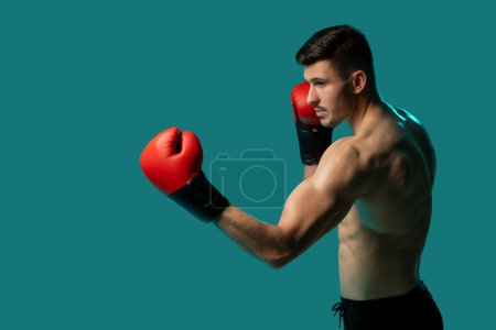 Un boxeador masculino, con guantes rojos y pantalones cortos negros, se está preparando para lanzar un puñetazo. Se encuentra en una posición de lucha, con un intenso enfoque en sus ojos contra un sólido fondo verde azulado.
