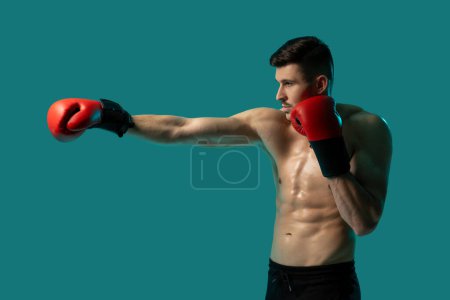 Photo for A muscular man is practicing boxing by throwing punches indoors against a solid blue background. He is wearing red boxing gloves and is shirtless, showcasing his toned physique and focused expression. - Royalty Free Image