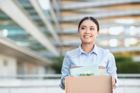 Foto de Excited Asian businesswoman is standing while holding a cardboard box. She appears focused and determined, possibly moving office or transporting items - Imagen libre de derechos