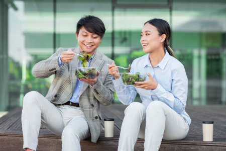 Two Asian individuals, dressed in business attire, are seen seated at a table. Both are actively engaged in consuming a bowl of salad, showcasing a healthy meal choice during their work break.