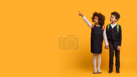 Ein afroamerikanisches Mädchen in Schuluniform mit Rucksack steht neben einem Jungen. Sie zeigt nach oben und schaut in diese Richtung, während der Junge mit neugieriger Miene zusieht.