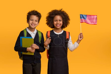 Afroamerikanische Jungen und Mädchen stehen stolz vor leuchtend gelbem Hintergrund, der Junge hält einen blauen Ordner in der Hand und trägt einen Rucksack, das Mädchen schwenkt die amerikanische Flagge.