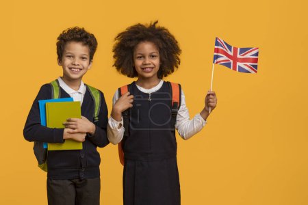 Two cheerful African American young students, a boy and a girl, stand against a vibrant yellow backdrop, proudly displays the Union Jack flag.