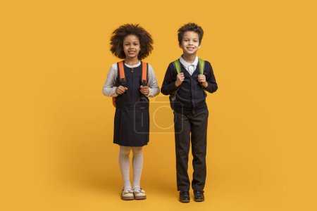 Zwei afroamerikanische Schulkinder mit Rucksäcken stehen vor einem leuchtend gelben Hintergrund. Sie scheinen bereit für einen Tag des Lernens, mit Büchern und Vorräten in der Tasche.