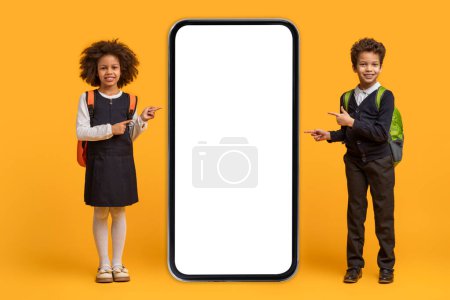 Foto de Dos jóvenes niños afroamericanos escolares, un niño y una niña, apuntan enérgicamente a una pantalla en blanco juntos contra un fondo naranja vibrante - Imagen libre de derechos