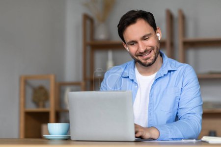 Un hombre alegre está participando en una videollamada, usando auriculares y sentado en un escritorio, sonríe cálidamente mientras mira la pantalla de su computadora portátil en un escritorio de madera en una habitación con iluminación suave y natural