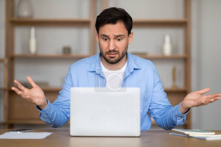 Ein Mann in einem lässigen blauen Hemd sitzt an einem Schreibtisch und zeigt einen verwirrten Gesichtsausdruck mit unsicher erhobenen Armen, während er auf seinen Laptop-Bildschirm blickt, der wahrscheinlich mit einer verwirrenden Arbeitssituation zu tun hat.