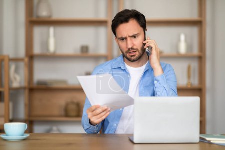 Photo for A man appears engaged in a serious conversation on his cellphone, simultaneously examining a document with a furrowed brow. The scene takes place in a well-lit home office - Royalty Free Image