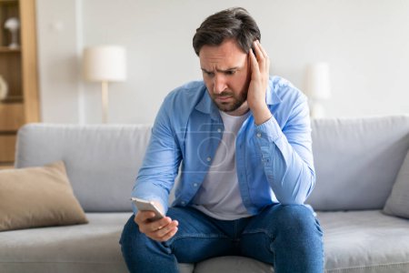 Photo for A man appears troubled or exasperated, sitting with a slouched posture on a grey couch. He cradles his head in one hand, with a smartphone held in the other. His expression conveys distress or concern - Royalty Free Image
