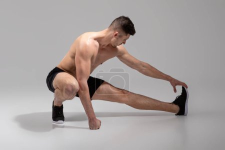 A fit, young man is seen stretching his leg muscles in a spacious, minimalist indoor gym, wearing black shorts and athletic shoes, practicing a deep lateral stretch to prepare for his workout routine