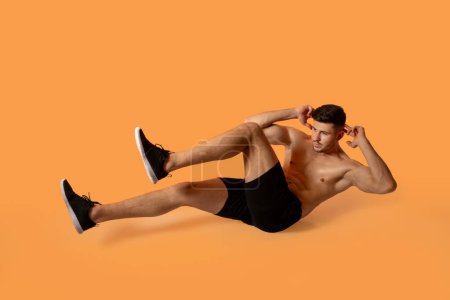 A fit man in black shorts and sneakers is performing a bicycle crunch exercise on an orange studio background. He is focused and demonstrating proper form.