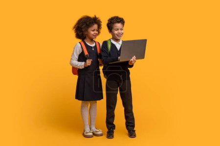 Un garçon et une fille afro-américains, vêtus d'uniformes scolaires intelligents avec des sacs à dos soignés, se tiennent côte à côte avec le garçon tenant un ordinateur portable ouvert. Ils regardent tous les deux l'écran