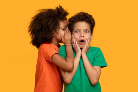 Two African American children are standing closely together, whispering to each other with animated expressions on their faces. The vibrant orange background makes their interaction stand out.