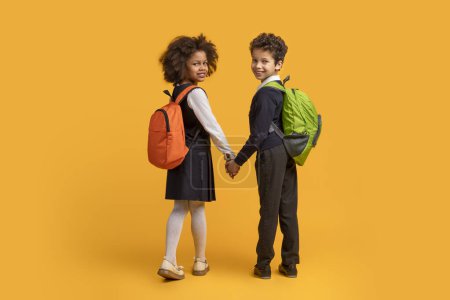 Foto de Dos niños afroamericanos, con mochilas, están tomados de la mano sobre un fondo amarillo brillante. Parecen estar listos para un viaje o aventura, retrospectiva - Imagen libre de derechos