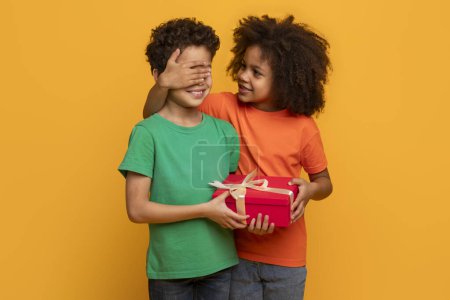 A young African American girl with curly hair is delightfully surprising a boy by covering his eyes with her hand while giving him a red gift box tied with a white ribbon