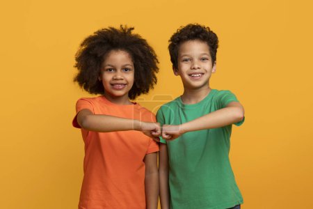 Deux jeunes frères et s?urs afro-américains joyeux se tiennent côte à côte sur un fond jaune vif, portant des t-shirts brillants et de couleur unie et souriant chaleureusement alors qu'ils joignent leurs poings ensemble