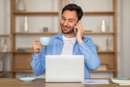 Ein Mann führt ein Telefongespräch, während er eine Tasse Kaffee in der anderen Hand hält. Er scheint Multitasking zu betreiben, balanciert das Telefon gegen sein Ohr, während er an der dampfenden Tasse nippt..