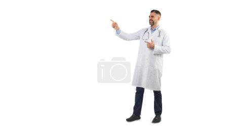 A man doctor is standing against a plain white background while pointing to the side and smiling. He is wearing a white lab coat with a stethoscope around his neck, copy space
