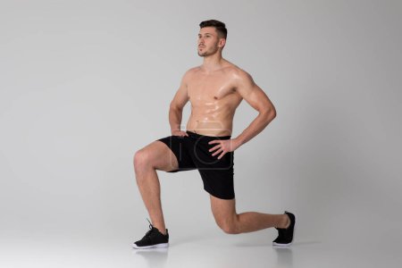 A man is exercising by performing squats on a gray background. He is bending his knees and lowering his hips, then pushing back up to a standing position