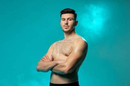 Un jeune homme confiant se tient torse nu, les bras croisés, regardant directement la caméra. Le fond est un bleu vif, accentué par des effets de brouillard, créant une atmosphère dramatique et frappante.
