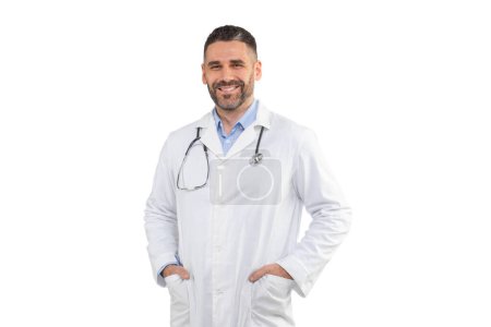 Foto de Un médico sonriente se para con confianza con las manos en los bolsillos, con una bata blanca y un estetoscopio, sobre un fondo blanco liso en un estudio. - Imagen libre de derechos