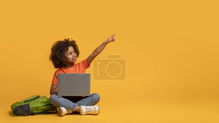 Ein junges afroamerikanisches Mädchen sitzt auf dem Boden, vor sich einen Laptop. Sie zeigt auf etwas auf dem Bildschirm und konzentriert sich intensiv auf die digitalen Inhalte, die angezeigt werden..