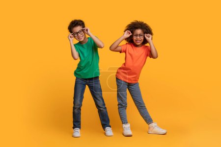 Afroamerikanische Jungen und Mädchen stehen Seite an Seite vor leuchtend gelbem Hintergrund, justieren spielerisch die Brille, machen Gesichter und genießen gemeinsam einen unbeschwerten Moment.