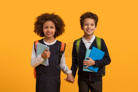 Two African American school children, a boy and a girl, stand hand in hand in front of a bright yellow background. The children are smiling and looking towards the camera