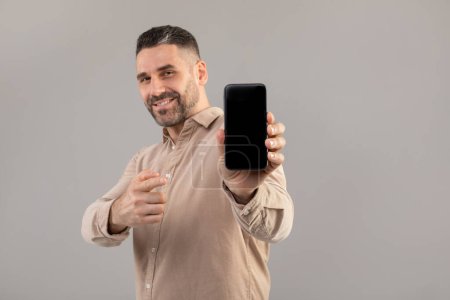 A bearded man in a beige shirt smiles while holding up a smartphone with a blank screen. The background is solid gray, and the man points towards the camera with his other hand, mockup