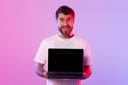 Foto de A cheerful young man with a beard is holding a laptop with a black screen, standing against a vibrant, colorfully lit background. - Imagen libre de derechos