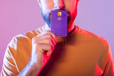 Foto de Un hombre con una camisa naranja sostiene una tarjeta de crédito púrpura cerca de su cara en un ambiente interior débilmente iluminado. El fondo presenta una mezcla de iluminación rosa y púrpura, creando una atmósfera vibrante. - Imagen libre de derechos
