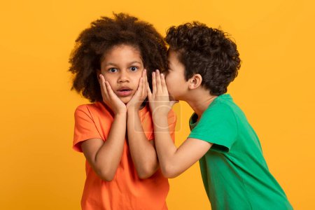 A young African American boy in a green shirt is whispering into the ear of his surprised sister, who is wearing an orange shirt. Her hands are placed on her cheeks, and her expression is astonishment