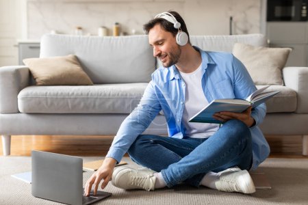 A cheerful man sits comfortably on the floor against a couch, wearing headphones and a blue shirt, engrossed in a book while simultaneously working or studying on a laptop