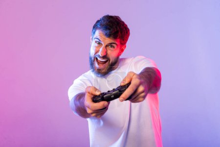 Foto de Un hombre barbudo juega intensamente a videojuegos, agarrando un controlador y mostrando una expresión excitada, iluminada por una vibrante iluminación de estudio rosa y azul.. - Imagen libre de derechos