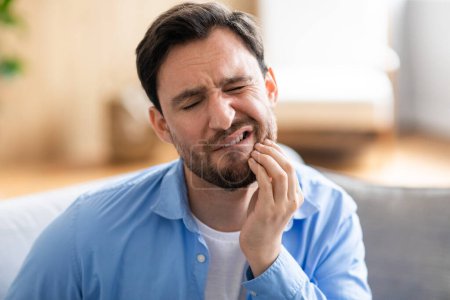 Un homme est montré assis sur un canapé, visiblement dans l'inconfort, tenant sa mâchoire avec une expression douloureuse due à un mal de dents. Il semble chercher un soulagement ou envisager de demander de l'aide dentaire.