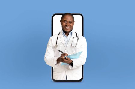 Foto de Un médico negro alegre vestido con una bata blanca con un estetoscopio alrededor del cuello, sosteniendo un portapapeles, parece emerger de una pantalla de teléfono inteligente colocada sobre un fondo azul claro. - Imagen libre de derechos