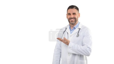 A man doctor, wearing a white lab coat and stethoscope, smiles as he gestures to the side. Background is a plain, well-lit studio setting, highlighting his professional attire, copy space