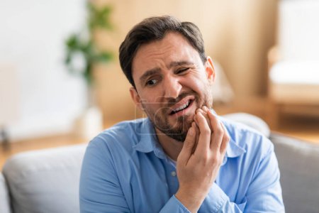 Ein Mann mit Unbehagen sitzt auf einer Couch und hält sein Gesicht vor Schmerzen aufgrund von Zahnschmerzen. Sein Gesichtsausdruck verrät sein Leiden, während er versucht, den Schmerz zu lindern.
