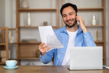 Photo for A man is sitting at desk while talking on the phone, holding a piece of paper in his hand. He appears focused as he communicates using the phone and glances at the paper intermittently. - Royalty Free Image