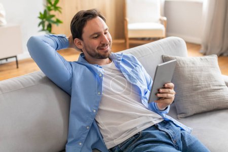 Foto de Un hombre está sentado casualmente en un sofá, absorto en el uso de un dispositivo de tableta. Aparece enfocado mientras interactúa con la pantalla, mostrando la tecnología moderna en la vida cotidiana. - Imagen libre de derechos