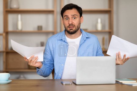 Un homme avec une expression surprise est assis à un bureau dans un bureau à la maison, tenant des papiers dans ses mains. Un ordinateur portable est ouvert devant lui, suggérant un moment de nouvelles inattendues ou de choc lié au travail.