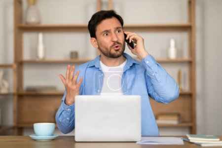 A man appears troubled or confused as he talks on his mobile phone with one hand gesturing, sitting in front of an open laptop, possibly working from home or an office
