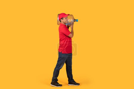 Drlivery hombre en una camisa roja y gorra que lleva un gran galón de agua sobre su hombro mientras está de pie sobre un fondo amarillo brillante sólido.