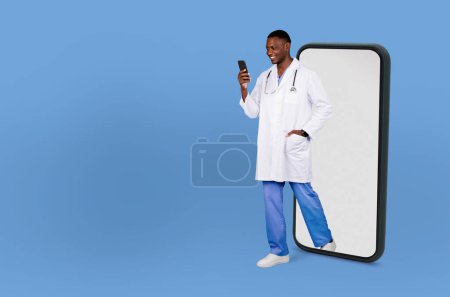 Un hombre negro profesional de la salud sonriente, vestido con una bata de laboratorio blanca y uniformes azules, camina a través de una pantalla gigante de un teléfono inteligente mientras mira su teléfono celular.